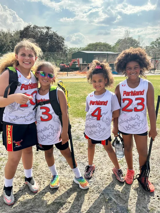 Four smiling Redhawks girls in white Parkland jerseys hold their lacrosse sticks