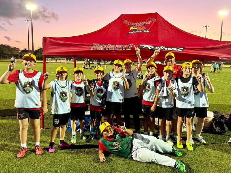 Young Redhawks players in white pinnies cheer under the red tent at dusk